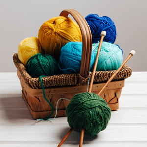 A brown wicker basket filled with blue and yellow balls of yarn. A green ball of yarn with two knitting needles sits in front of the basket. 