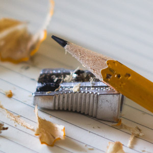 A sheet of lined paper with a pencil sharpener and sharpened pencil laying on top of it. Pencils shavings are scattered around the items.
