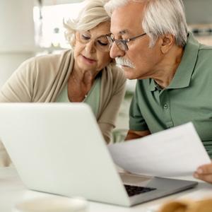 An older couple holding paperwork and reviewing information on a laptop screen. 