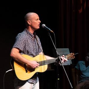 Dave Martin plays guitar on stage, lit by spotlight in a dark room