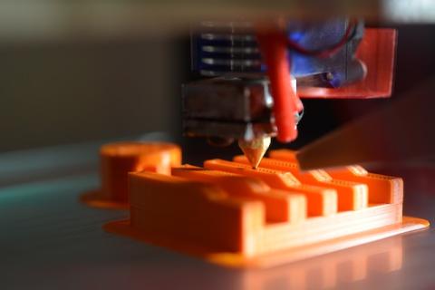 A 3D printing extruder is extruding orange filament onto a printing bed. This photo is by Gavin Allanwood. 